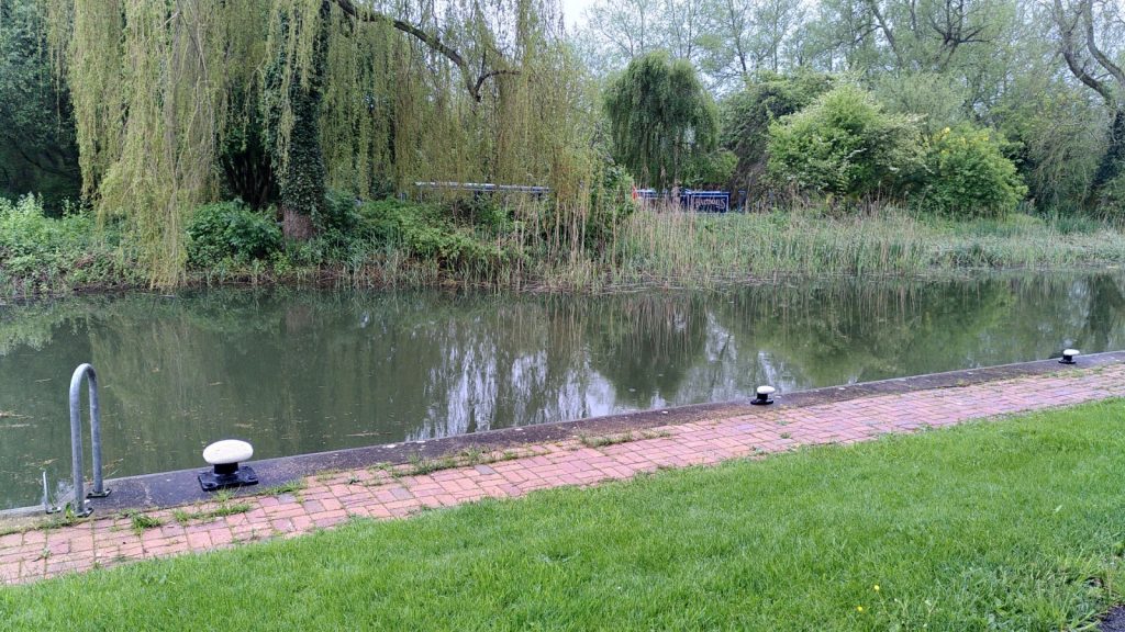 View across a canal.  On the near side there are a few bollards for mooring up. The opposite bank is obscured by tree foliage.  A narrowboat is just visible through the leaves.