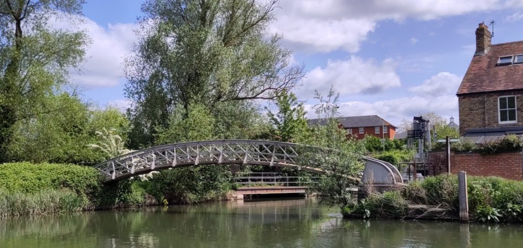 View across the river in to a side channel.  A grey metal bridge arches low over the entrance to the side channel.  Under it the channel has a metal fence along its side.