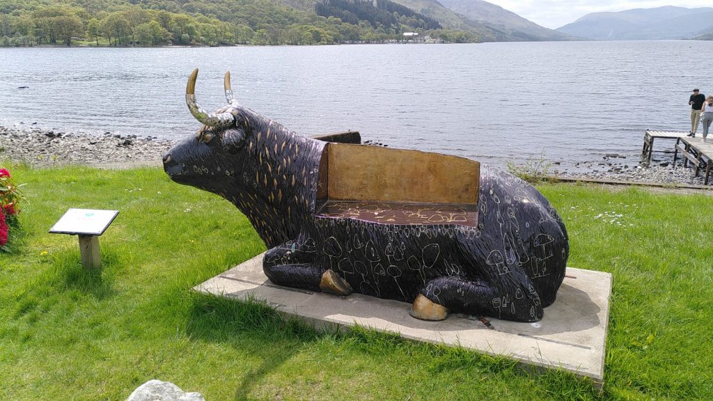 A statue of a highland cow lying down on a plinth.  The back of the cow has a segment removed so that it can be used as a bench.  Beyond the cow is a view over Loch Earn.