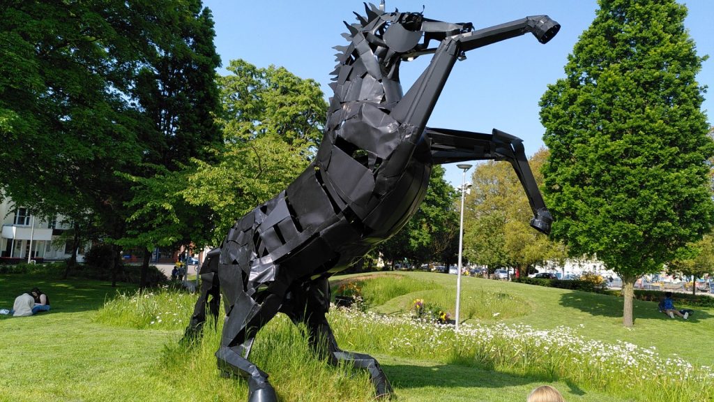 Statue of a black horse standing on its hind legs flailing its front legs.  The statue is life-size.  The setting is a park with trees.  The sky is bright blue and cloudless.
