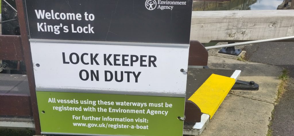 Sign on the lock gate saying Welcome to King's Lock. Underneath are two removable panels.  The upper one has a white background and says Lock Keeper On Duty.  The lower board is green with a longer message on it.