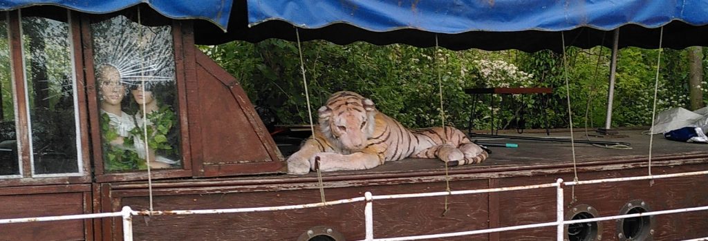 A life-size tiger statue lying on a boat.  Human faces can be seen peering out of an adjacent window.  They are statues too.