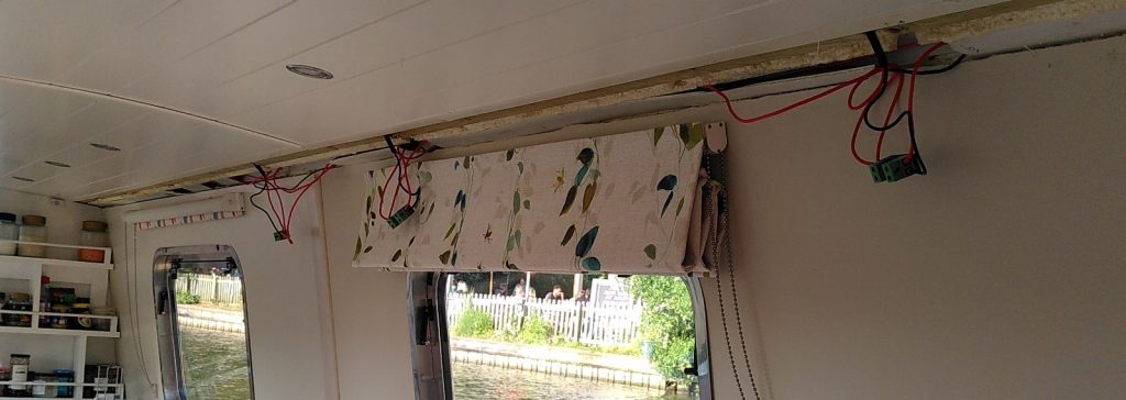 Inside the saloon of a narrowboat.  The white painted wooden trim along one edge of the ceiling has been removed.  Dangling from the ducting are wires.  Three  green diode blocks hang from the wires