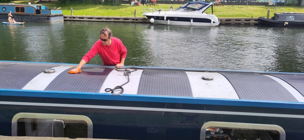 The roof of a narrowboat is covered with solar panels.  A man is standing on the gunwale on the far side of the boat wiping the panels with a cloth.  Behind him other boats can be seen moored on the other bank of the river.