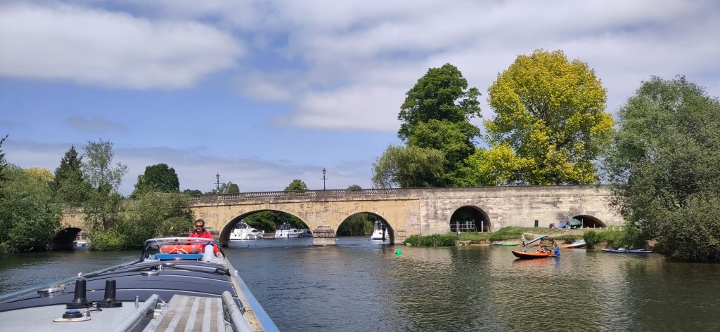 A multi-arched stone road bridge can be seen from the front of a narrowboat that has just come under it.  Beside the minor arches of the bridge are bathers, canoes and paddles boards.  The blue sky has some light clouds.