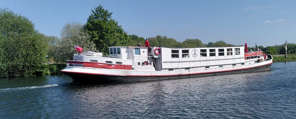 A large river boat heading downstream.  The crew visibe on board make it possible to judge the scale of the boat.  It is probably twice as high as a narrowboat, more than twice as wide and significantly longer.