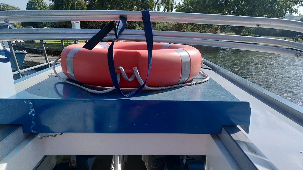A fluorescent orange life ring sitting on top of the blue rear hatch of a narrowboat.  Pram cover poles are propped up by a plastic bottle so that they are above the ring.