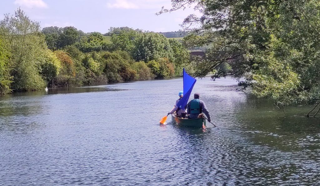 Two people in a canoe on a wide tree-lined river.  The person in front has what appears to be a makeshift sail on their back.