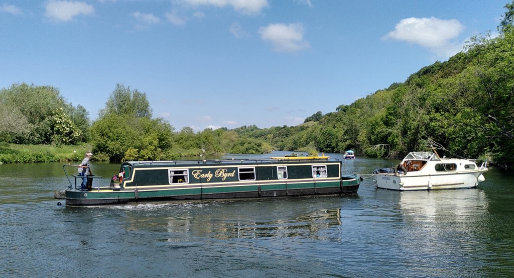 A white plastic boat towing a narrowboat across thee width of a wide river.  The banks are tree-lined and there are small fluffy clouds in the blue sky.