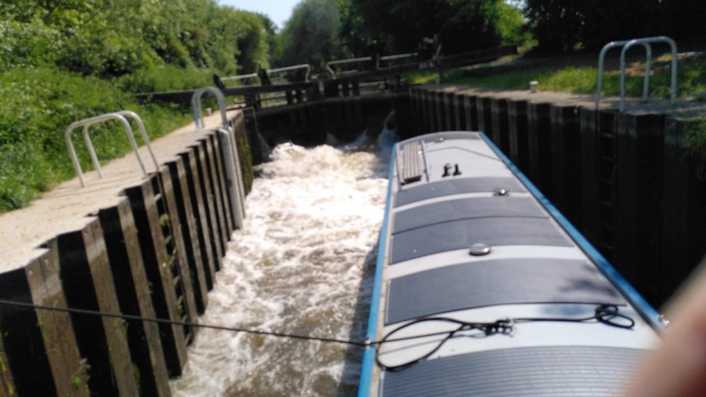 A narrowboat in a lock viewed from halfway along the roof.  A rope from the centre of the roof is fixed to one side of the lock.  The bow of the narrowboat is resting on the opposite side.   The water in the lock is churning and foamy.