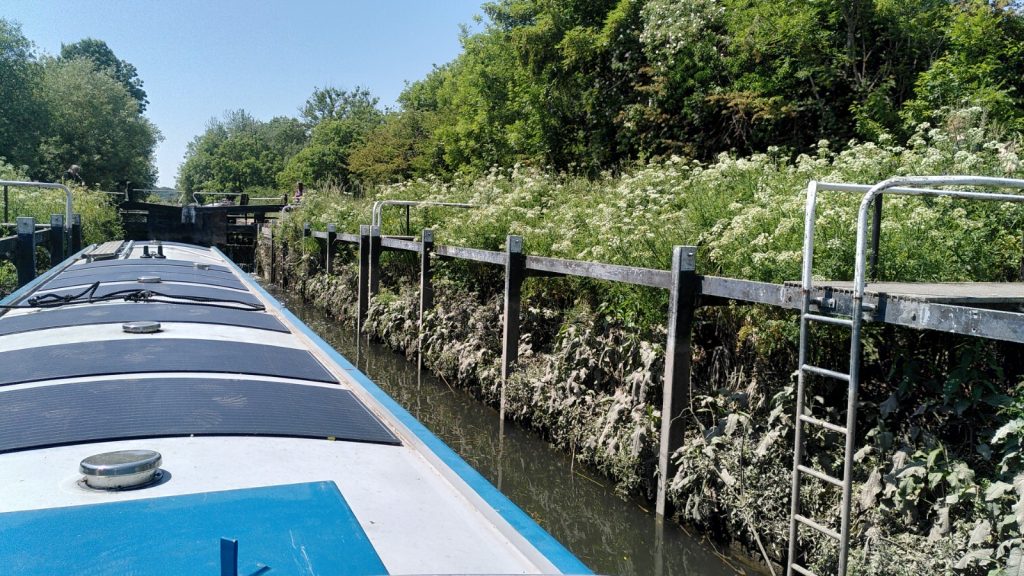 A narrowboat in an unusual lock.  The sides of the lock are a framework, behind which is a sloping bank covered in vegetation.