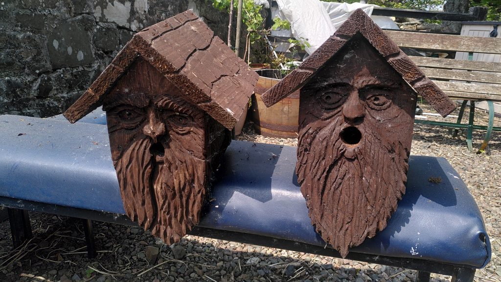 Two bird boxes sitting on a blue bench in the sunshine.  The wooden bird boxes have faces carved in their fronts.  The faces have long beards, with the mouth forming the circular entrance to the box.