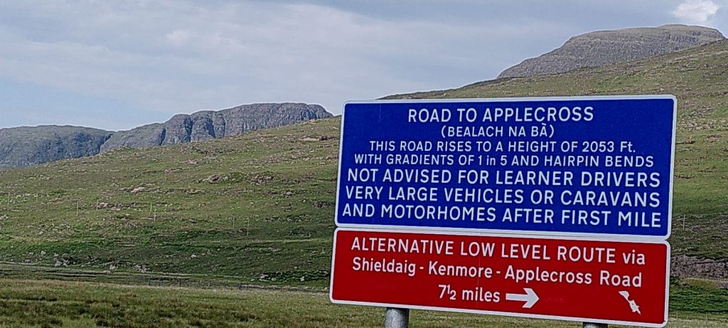 Road sign with mountains looming behind. The sign says: "Road to Applecross (Bealach na Bà). This road rises to a height of 2053 feet with gradients of 1 in 5 and hairpin bends. Not advised for learner drivers very large vehicles or caravans and motorhomes after first mile."