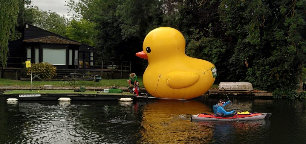 An inflated yellow rubber duck at the side of the river.  The duck looks like a standard bath toy, except that it is 12 feet high.  It is attached to a small boat on which is a golf green with flag, balls and a lawn mower.