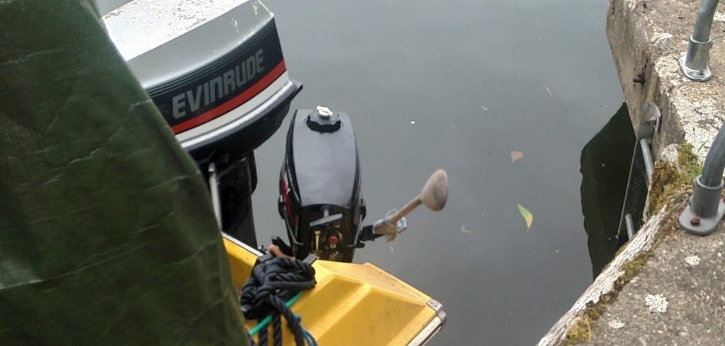 An outboard motor at the back of a small boat.  The handle of the motor has been extended with a wooden spoon.
