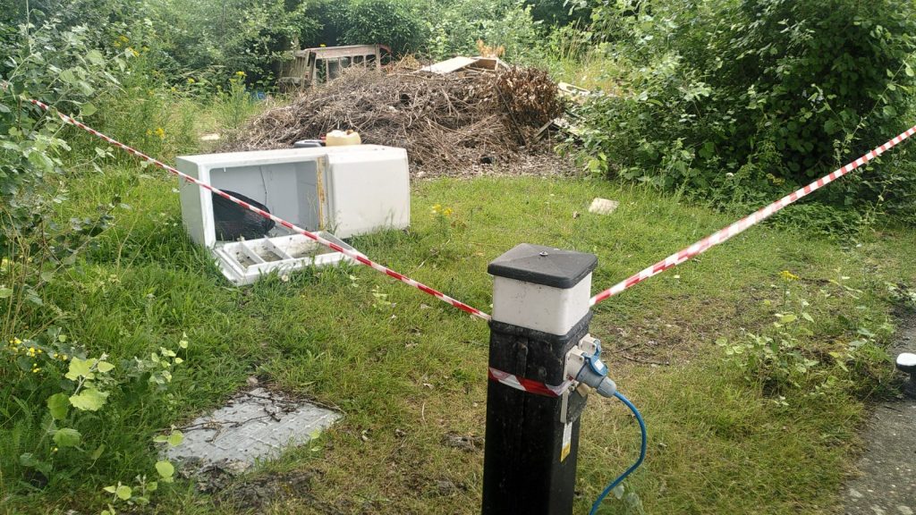 A short black post with a light at the top has an electric hook-up cable plugged in to it. Red and white hazard tape is wrapped around the post and extends away.  Behind the tape is a small rubbish dump.