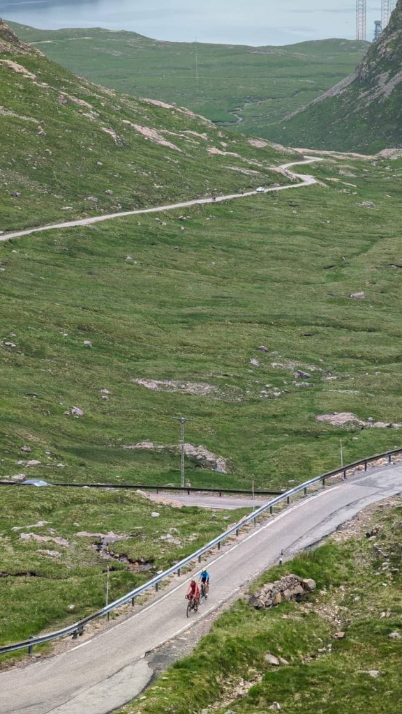 Looking back down the road from The Bealach.  Below are two cyclists, one in red and one in blue cycling up a stretch of road. Behind the road snakes under them to the other side of the valley and disappears towards the distant sea.