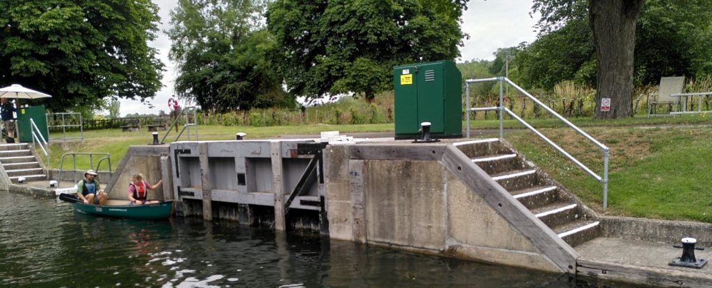 View across a lock on the River Thames.  The usual line of bollards is interrupted by an additional lock gate. Two people in a canoe are holding on the lock wall.