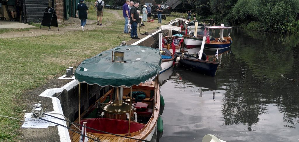 A collection of small steam powered boats tied up at a river wharf.  Each boat seats about four people and has a boiler and funnel.