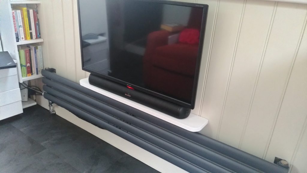 TV screen hangs from a  wall.  Below the TV is a wide low radiator.  Separating the TV from the radiator is a narrow wooden shelf on which a sound bar rests.