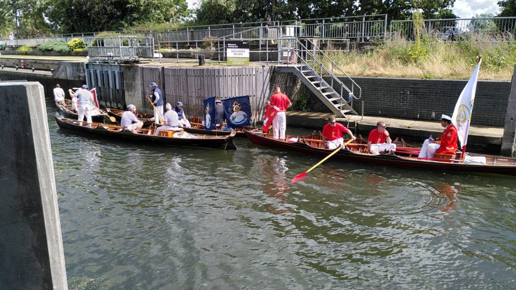 Several small wooden rowing boats are manoeuvred into a river lock.  Each boat has three or four men.  Each boat's occupants are dressed similarly, red jackets in one, blue in another and white in the third.