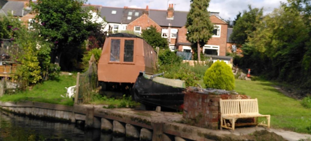 A repurposed narrowboat in a riverside garden.  The cabin section looks like it is in use as a shed, while the bow sits separately alongside as an ornament.