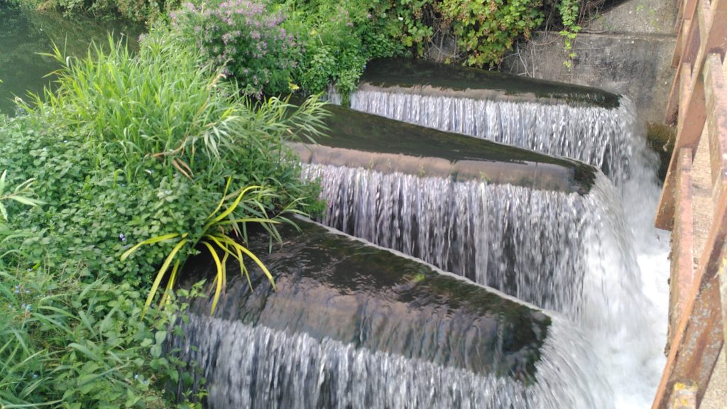 A weir on the River Kennet.  The water cascades over the edges of a wall arranged in a sharp zig-zag pattern.  The fingers of water are two or three times as long as they are wide.