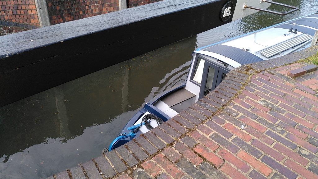 A narrowboat is visible between the arm of an open lock gate and the scalloped brick wall below it.  A metal frame over a foot square forms a handle to allow the arm to be pulled back without stepping over the edge.