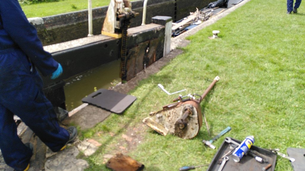 An open lock gate with part of the paddle winding mechanism sitting on the grass alongside.There is a mat at the lock edge for the worker to lie on, and various tools within reach.