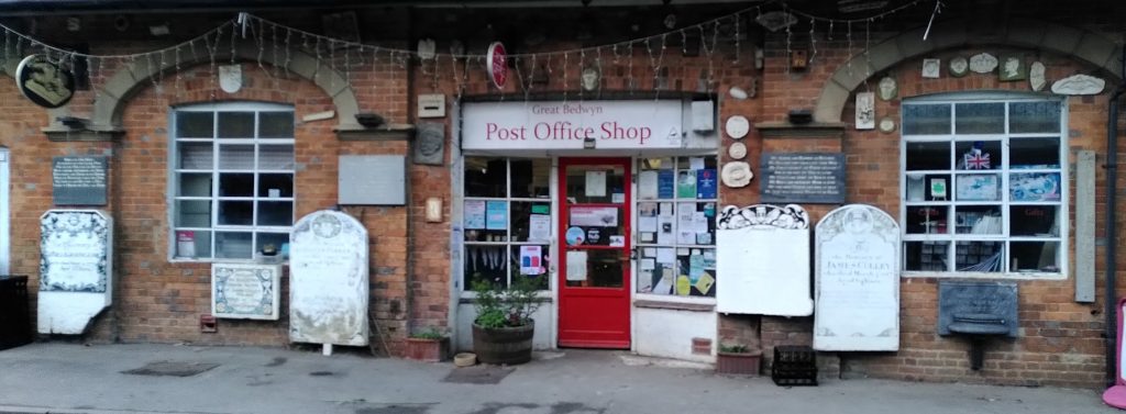 Great Bedwyn Post Office.  A small old fashioned shop entrance in a larger brick building.  Along the shop frontage are a number of tombstones and similar stone plaques.