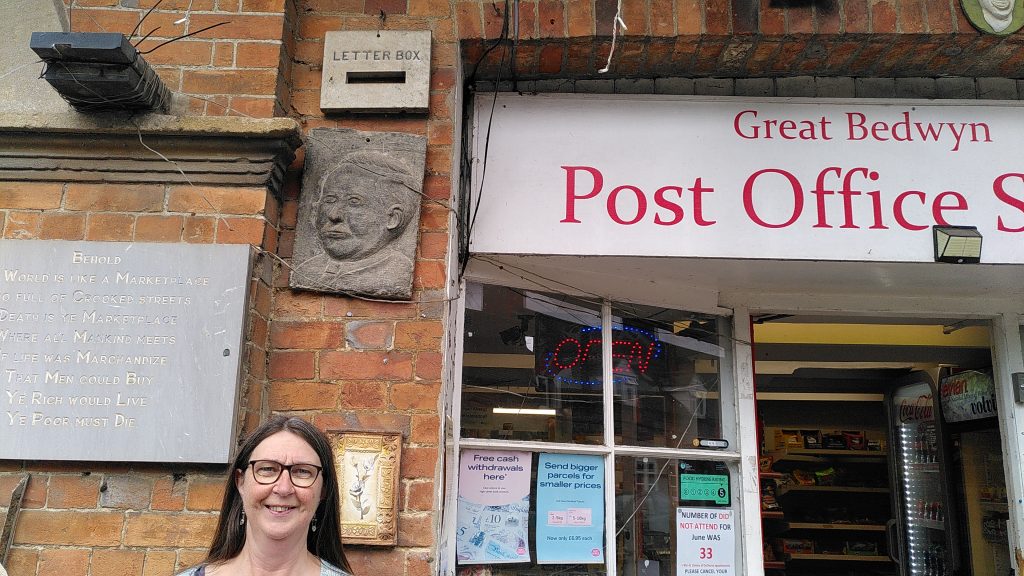 Clare standing outside the Great Bedwyn Post Office.  The window extends well above her head and has the main Post Office sign above it.  Well out of reach, and level with the top of the sign is a stone plaque saying "Letter Box" with a slot.
