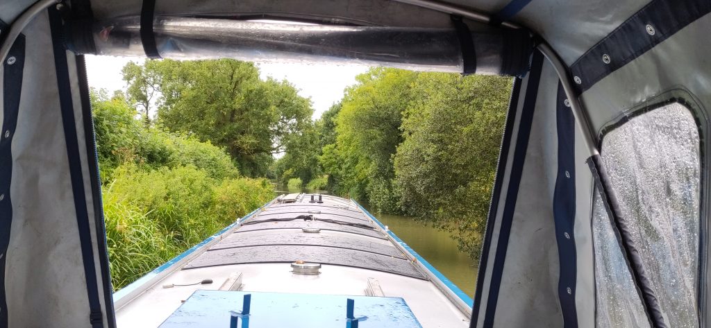 View from the steering position of a narrowboat with the pram cover up.  The foreground is the roof of the boat.  On both sides trees are visible along the canal edge, a small amount of water is visible at the side