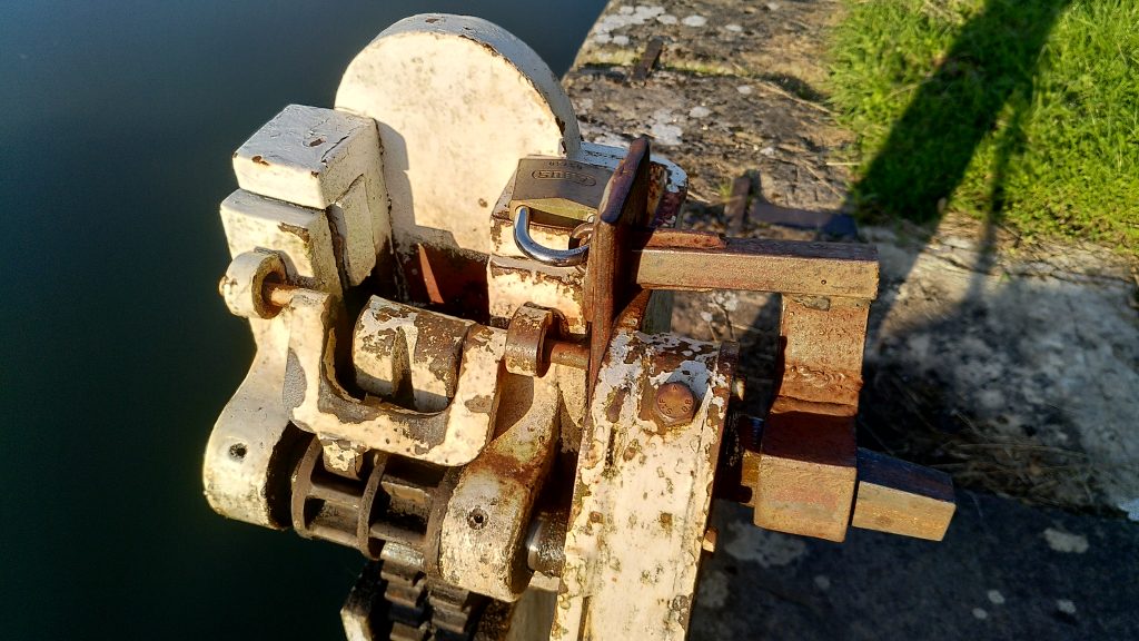 A close-up of the gear mechanism for aground paddle.  The white painted mechanism is disabled by a collar on the spline held in place by a brass padlock.