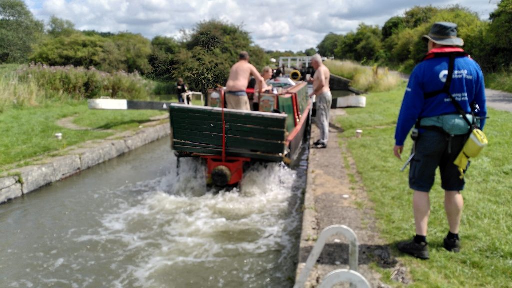 A paddle-powered narrowboat exiting a lock.  The driver and a companion on the bank are shirtless.  A lock-keeper is standing watching them - in awe or bewiderment.