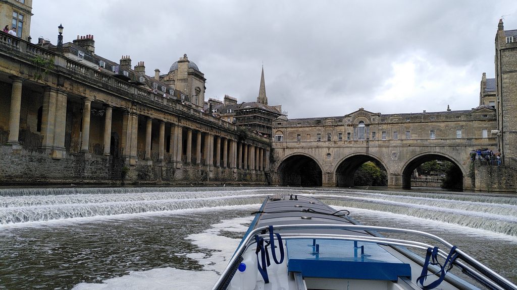 View from the helm of a narrowboat.  The boat is approaching a horseshoe shaped weir from below.  Above the weir, Pulteney Bridge crosses the river.  A riverside walk has fine stone columns supporting the pavement above.