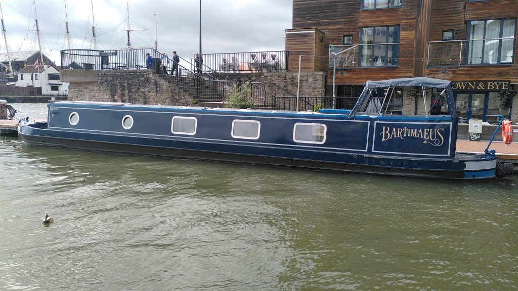 Bartimaeus moored in Bristol Floating Harbour. There is a stone harbour  wall behind the boat, and beyond it the masts of the SS Great Britain across the harbour.