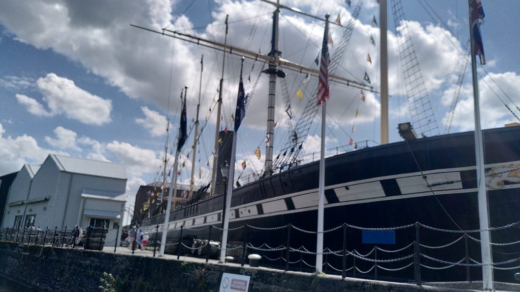 Looking up at the SS Great Britain from the water.  A stone wall separates us from the ship.  Its superstructure towers over us and the masts are considerably higher again.  Colourful flags give a message.