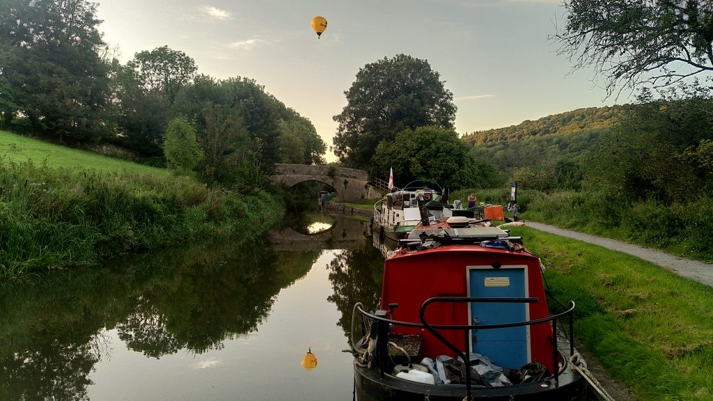 A view along a canal to a stone humped back bridge.  A hot-air balloon is directly above the bridge and reflected in the canal below it.