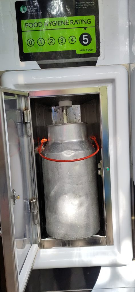 Milk being dispensed in to a small metal milk churn. The churn is sitting inside a vending machine.  A sign on the machine shows a hygiene rating of 5 (the best).