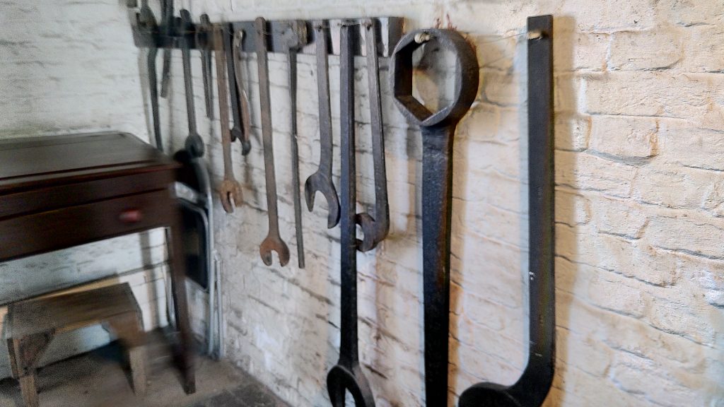 A set of spanners and other tools hanging from a white-washed wall.  The tools are on a huge scale.  Most are several feet long, some are five feeet long or more.
