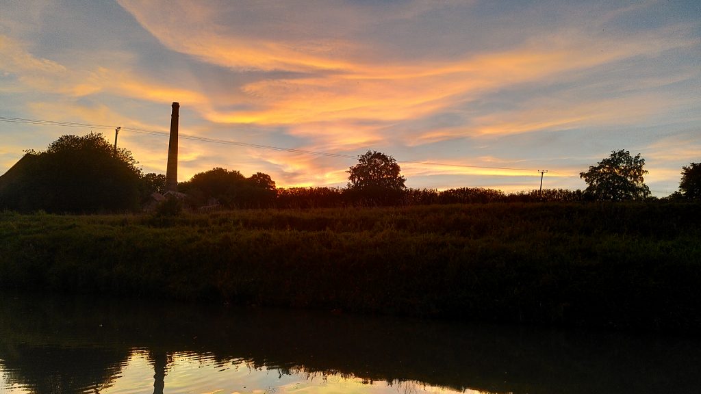 Multicoloured sky.  A tall chimney interrupts the skyline. The sky is reflected in the canal.