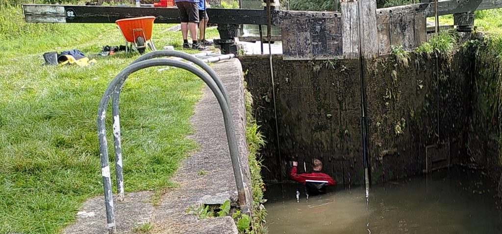 Worker repairing lock gate.  A view in to a lock.  The water is low but the gates are closed.  A man is standing in the water looking at the holes in the gate.  The feet of his companions can be seen on the lock side near the gate arm.