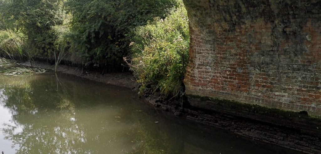 Reduced water level in a canal.  The view is from under the arch of a bridge.  The stonework on the far side is wet above the water line for two feet or so.  The bank further along the canal also looks low.  Some reeds are lying flat on the surface.