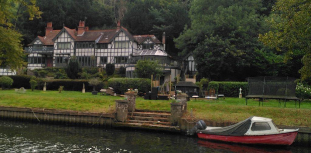 Large riverside house with black and white timbered walls.  The house is extensive and set back from the river in its own grounds.  There is a small boat moored on the river.
