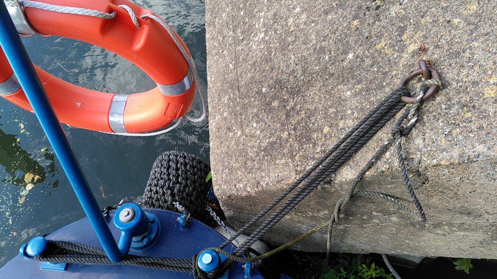 Looking down from the stern deck of a narrowboat.  The stern is separated from a concrete block by a small plastic fender.  The rear fender of the boat is also resting on the concrete.  The boat is tied to a ring attached to the concrete.