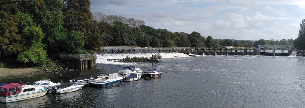 View of Teddington Lock Weir from downstream. In the foreground some moored boats sit in low water levels.  Behind them a curtain of white water shows the location of the weir.  Just visible through the railings at its top are a row of boats waiting to use the lock.