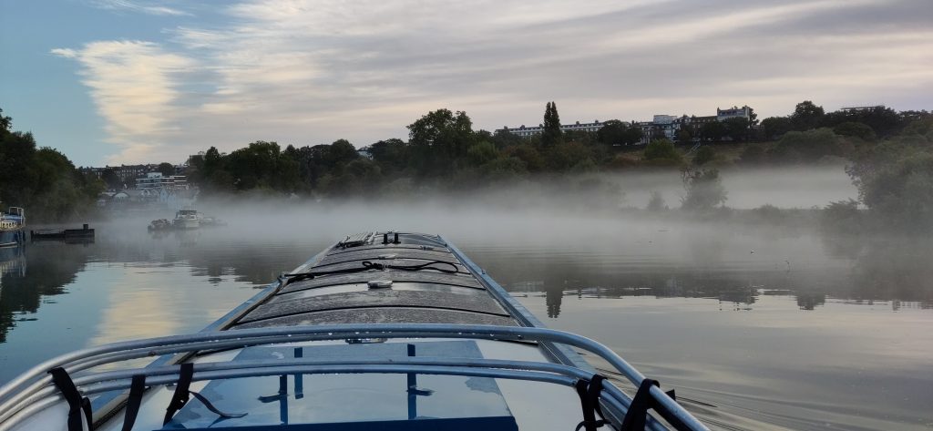 View from the helm of a narrowboat.  In front of the boat a bank of mist sits low on the water.  Behind, the side of the valley is mostly wooded.  Light cloud is giving way to blue sky.