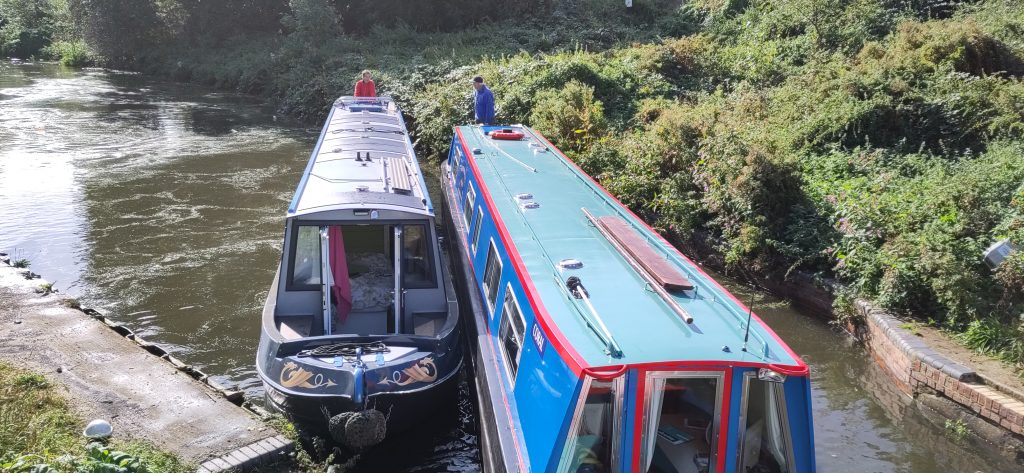 Two narrowboats diagonally across the canal.  The drivers of each are in the vegetation on the far bank.