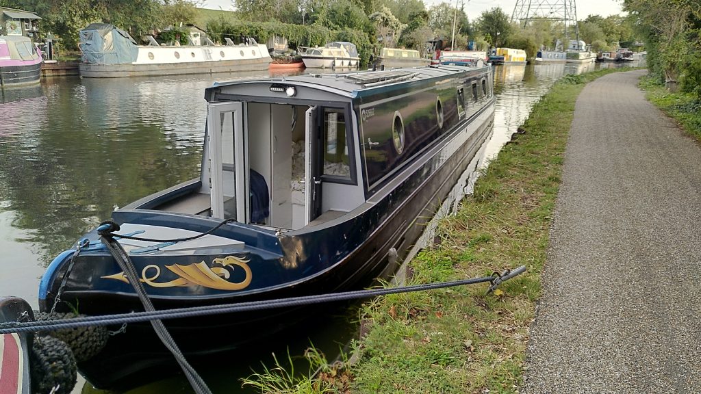 Narrowboat moored at a jaunty angle.  The front of the boat looks quite close to the bank, but there is a visible gap at the back, even though it is further away.