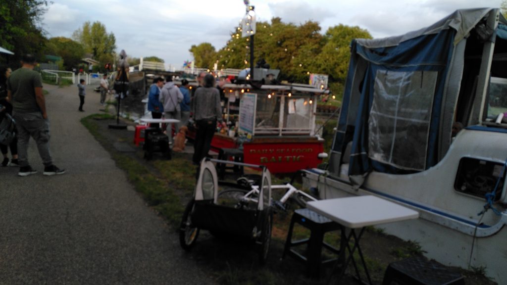 Fish take away boat.  A boat that looks like a trawler decked out with fairy lights is acting as a fish bar.  A number of people are gathered around making and collecting their orders.  There are a number of tables and chairs arranged either side of the towpath for customers use.